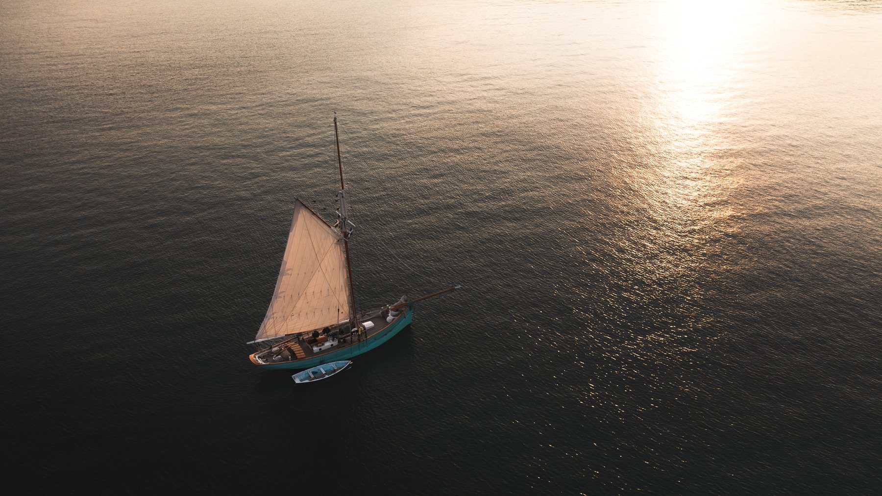 Aerial view of a classic sailboat under sail at sunrise, towing a small tender